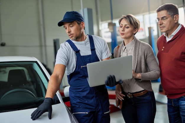 Riparatore automobilistico e coppia che esaminano i problemi dell'auto durante l'utilizzo del laptop in un'officina