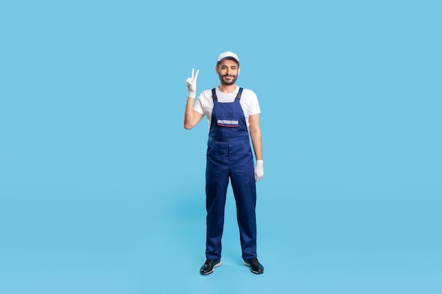 Riparatore esperto felice a figura intera in tuta e guanti protettivi in piedi, mostrando il gesto di vittoria e sorridendo soddisfatto. Professione del terziario, manutenzione della casa. girato in studio isolato