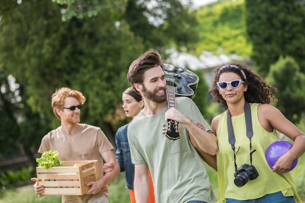 Riposa, sulla natura. Giovani ragazzi amichevoli con cibo e chitarra con ragazze carine che vanno energicamente a fare un picnic parlando di buon umore