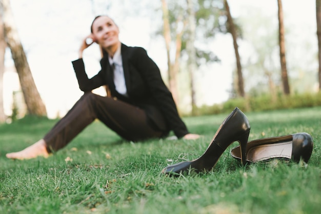 Riposo dopo il lavoro in ufficio Bella giovane donna d'affari in abito nero è seduta nel parco sul prato e sorridente Scarpe da donna in primo piano