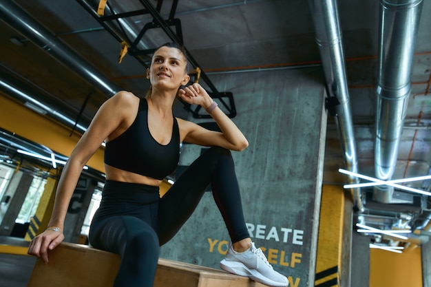 Riposo dopo l'allenamento. Giovane bella ragazza atletica in abiti sportivi che si siede sulla scatola di salto in legno crossfit in palestra e guardando da parte. Persone sportive, allenamento, concetto di stile di vita sano