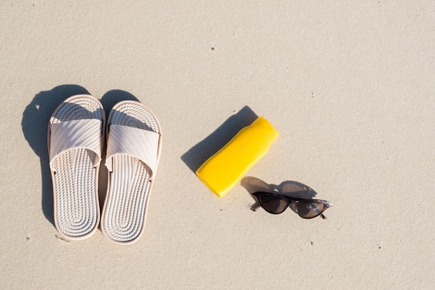 Riposo sulla spiaggia: pantofole, crema protettiva e occhiali da sole su sabbia pulita. Vista dall'alto di accessori per vacanze al mare o vacanze estive