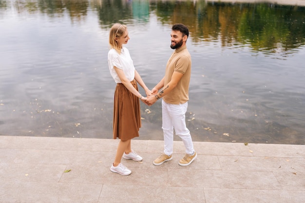 Ripresa a tutto campo di allegre giovani coppie innamorate in piedi che si tengono per mano vicino al bellissimo lago della città che sorride guardandosi l'un l'altro il giorno dell'estate