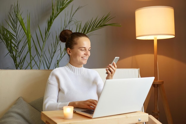 Ripresa al coperto di una bella donna impegnata con l'acconciatura da chignon che indossa un maglione bianco seduta sulla tosse e lavora al computer portatile e usa il cellulare guardando il display del cellulare con un sorriso