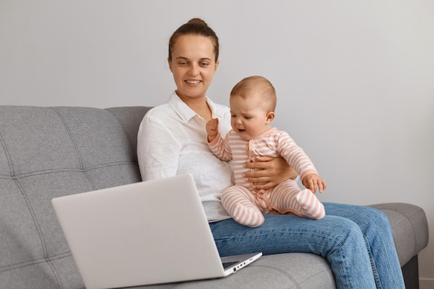 Ripresa al coperto di una donna soddisfatta che indossa camicia bianca e jeans seduta sul divano al coperto e tiene in braccio il bambino, guarda lo schermo del laptop, guarda un film o fa una videochiamata.