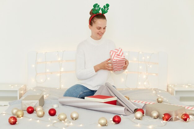 Ripresa al coperto di una donna sorridente che si prepara per le vacanze di Natale, avvolgendo la scatola regalo mentre è seduta sul letto in camera da letto bianca, indossando abiti in stile casual e oggetti di scena per le feste.