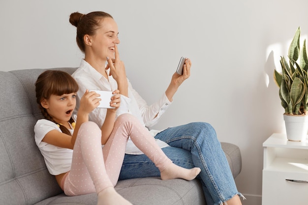 Ripresa al coperto di una donna sorridente positiva che indossa camicia bianca e jeans seduta sul divano con sua figlia e parla tramite videochiamata, puntando il dito verso l'alto, raccontando la sua nuova idea.