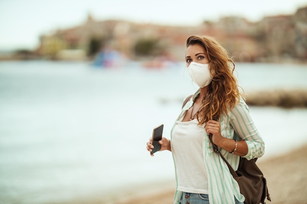 Ripresa di una giovane donna attraente con maschera protettiva N95 che si gode una vacanza sulla spiaggia durante il COVID-19.