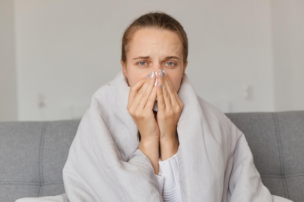 Ripresa in interni di donna malata con mal di testa seduta sotto la coperta e con in mano un fazzoletto che soffre di naso che cola guardando la fotocamera con espressione triste e sguardo malsano