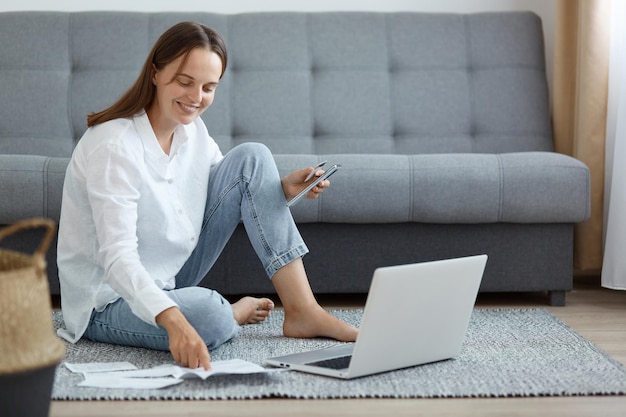 Ripresa in interni di donna sorridente che indossa una camicia bianca e jeans seduta sul pavimento vicino al divano utilizzando un laptop in possesso di carta di credito che controlla le informazioni nelle bollette su carta