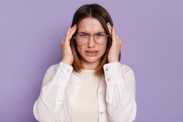 Ripresa in interni di frustrata donna dai capelli scuri malsana che indossa una camicia bianca in posa isolata su sfondo viola accigliata massaggi viso templi che soffrono di terribile mal di testa