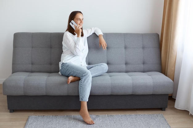 Ripresa in interni di giovane donna adulta con i capelli scuri che indossa una camicia bianca e jeans seduta sul pavimento e distoglie lo sguardo parlando al telefono con felice espressione facciale positiva