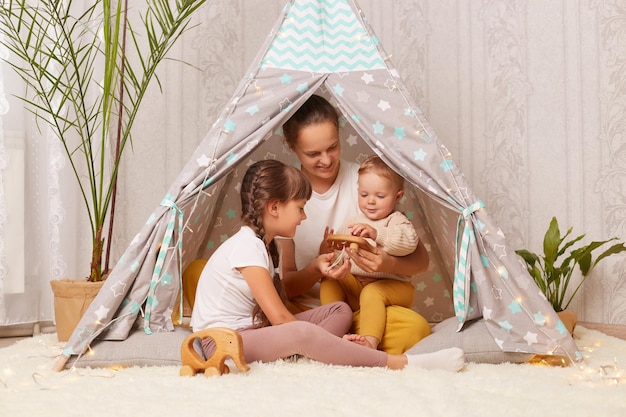 Ripresa in interni di madre con due bambini seduti insieme in wigwam teepee a casa mamma che gioca con bambini in età prescolare e infanti che mostrano un giocattolo ecologico in legno al suo bambino