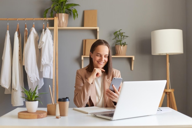 Ripresa in interni di sorridente attraente stilista donna dai capelli castani che indossa una giacca beige seduta sul posto di lavoro davanti al computer portatile e utilizza lo smartphone esprimendo emozioni positive