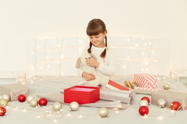 Ripresa in interni di una bambina seduta sul letto su una coperta spesso con un regalo in mano, che apre una scatola con un regalo di Natale, che posa in camera da letto decorata con atmosfera e ghirlande di Capodanno.