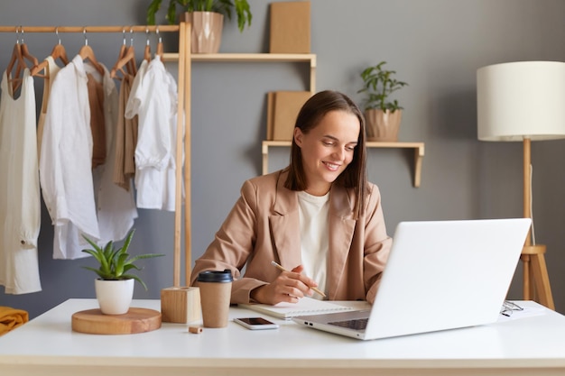 Ripresa in interni di una bellissima stilista donna felice che indossa una giacca beige seduta davanti al computer portatile con vestiti appesi su grucce sullo sfondo disegnando schizzi di nuovi abiti per la nuova collezione