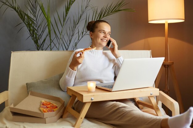 Ripresa in interni di una donna che si rilassa mentre trascorre il giorno libero a casa utilizzando il notebook mangiando pizza e parlando al cellulare conversando distogliendo lo sguardo con un sorriso