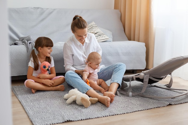 Ripresa in interni di una donna dai capelli scuri che indossa una camicia bianca e jeans seduta sul pavimento con le sue figlie che giocano con i bambini sul tappeto trascorrendo del tempo con i bambini