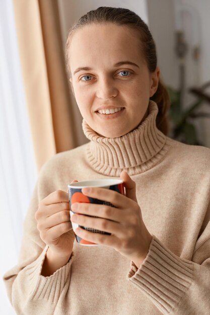 Ripresa in interni di una donna positiva sorridente con i capelli scuri che indossa un maglione beige in piedi con in mano una tazza di caffè e tè guardando la fotocamera con una piacevole espressione facciale