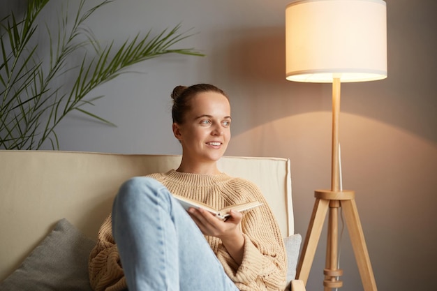 Ripresa in interni di una donna sorridente e soddisfatta che indossa un maglione beige e jeans seduta sulla tosse e distoglie lo sguardo tenendo in mano la lettura di un libro che sogna l'amore come nella trama di un romanzo