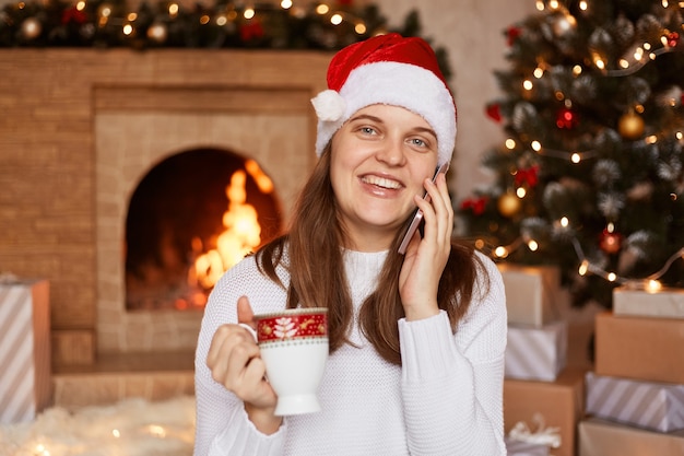 Ripresa in interni di una donna sorridente felice che indossa un maglione bianco e un cappello di babbo natale, seduta vicino al camino e all'albero di Natale, che guarda l'obbiettivo mentre si conversa tramite telefono cellulare.