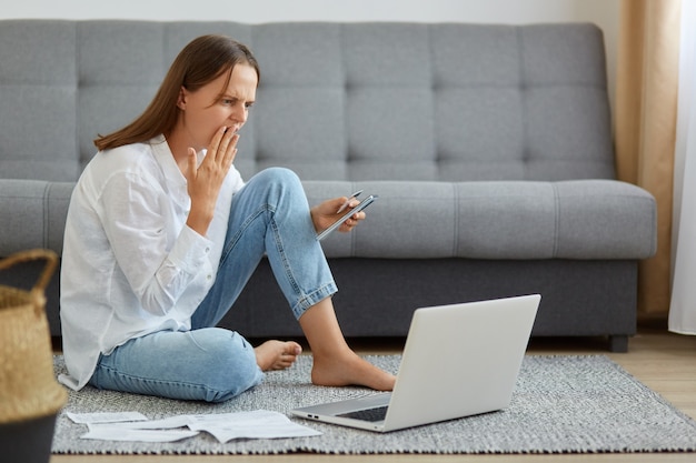 Ripresa in interni di una donna spaventata che indossa una camicia bianca e jeans, seduta sul pavimento con un computer portatile, guardando lo schermo del notebook, con in mano una carta di credito, inviando denaro nel posto sbagliato.