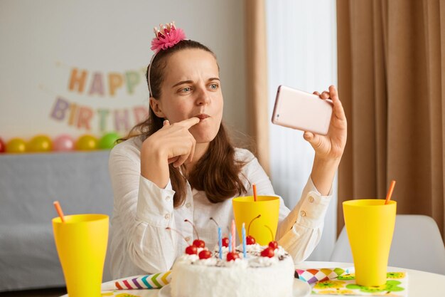 Ripresa in interni di una giovane donna caucasica che indossa una camicia bianca che festeggia il compleanno da sola guardando video sullo smartphone o facendo una videochiamata con congratulazioni che assaggiano la torta guardando lo schermo del telefono