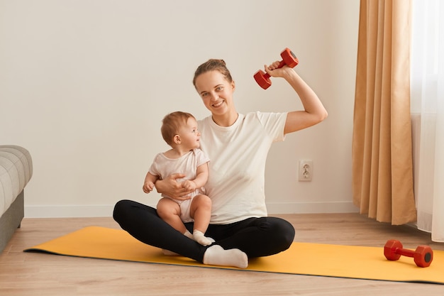 Ripresa interna della madre con la figlia del bambino che fa ginnastica e esercizi di fitness a casa seduta sul tappetino da yoga donna che tiene il bambino neonato e braccio sollevato con manubrio