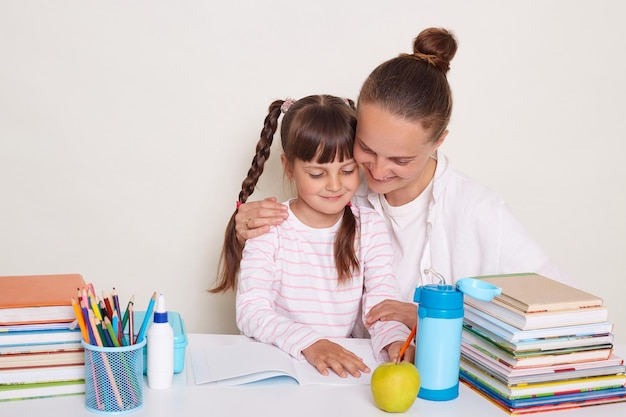 Ripresa interna di giovane donna con acconciatura da panino che fa i compiti con la famiglia della figlia che legge insieme madre che abbraccia la sua piccola scolaretta intelligente in posa isolata su sfondo bianco