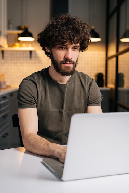 Ripresa verticale di un giovane libero professionista barbuto concentrato che usa la digitazione sulla tastiera del laptop scrivendo e-mail o un messaggio seduto al tavolo in cucina