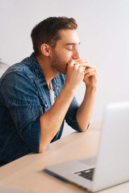 Ripresa verticale di un libero professionista affamato che mangia hamburger guardando video online sul computer portatile durante la pausa