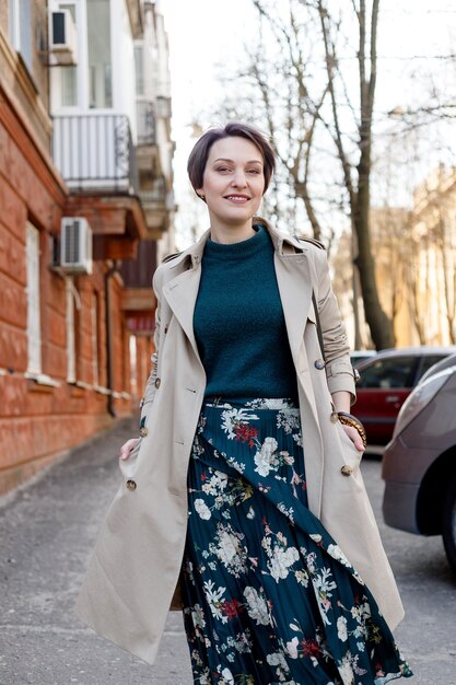 Riprese di ritratto di una donna attraente ed elegante, ragazza cammina in città all'aperto. Elegante immagine moderna e femminile, stile. Ragazza con un mantello o un cappotto beige e un vestito verde