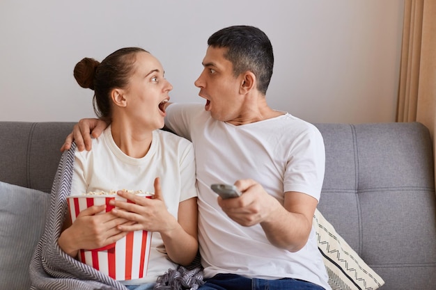 Riprese in interni di giovani coppie scosse che guardano film horror in tv e mangiano popcorn a casa seduti sul divano con la bocca aperta che si guardano con shock