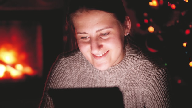Riprese toniche di una giovane donna sorridente che guarda video su tablet di notte in soggiorno con caminetto acceso