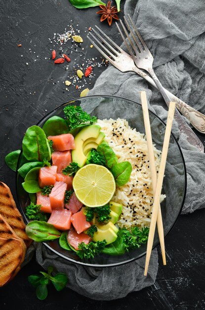 Riso con salmone, avocado e spinaci In un piatto nero su sfondo di legno Vista dall'alto Spazio libero per il testo Disposizione piatta