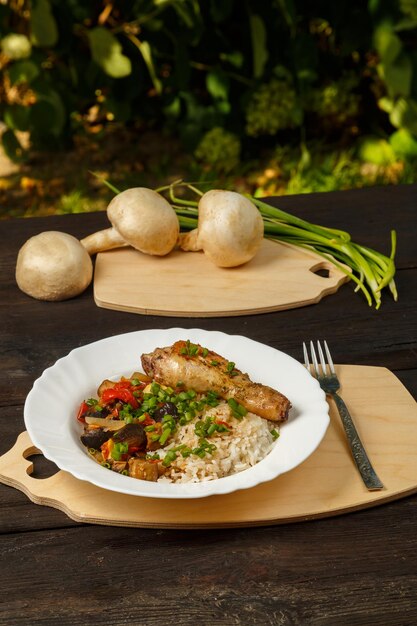 Riso con verdure stufate in salsa e coscia di pollo in un piatto bianco su un tavolo di legno accanto a champignon e cipolle verdi sul bordo