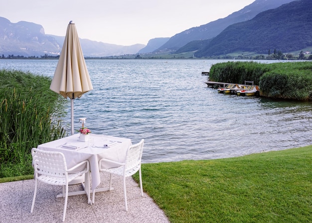 Ristorante di strada di lusso con tovaglia bianca e fiori al Lago di Caldaro in Alto Adige. Caffè italiano sul marciapiede con tavolo e sedia al Lago di Caldaro. Design della terrazza stile di vita per la cena.