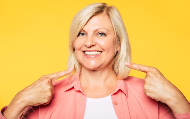 Risultato perfetto della chirurgia dentale. La bella donna anziana bionda felice sta indicando il suo sorriso sano a trentadue denti e guarda la telecamera