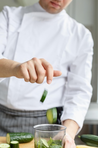 Ritaglia il colpo senza volto dello chef maschio in uniforme che mette pezzi di sedano tagliati nel contenuto per il frullato