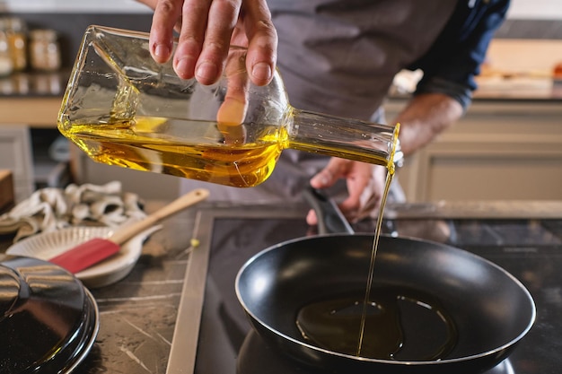 Ritaglia il cuoco maschio anonimo versando l'olio dalla bottiglia nella padella sul fornello mentre cucini al bancone in cucina leggera