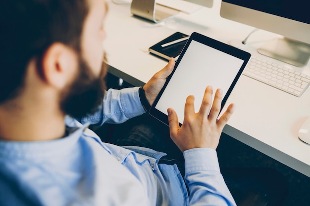 Ritaglia il ragazzo barbuto toccando lo schermo vuoto del tablet moderno mentre è seduto alla scrivania in ufficio moderno. Impiegato anonimo utilizzando tablet