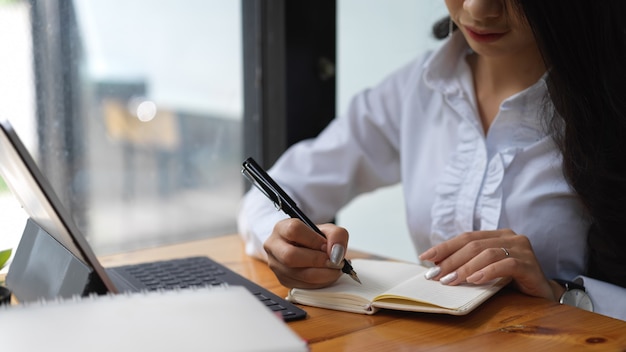 Ritagliata colpo di donna che lavora con tavoletta digitale e scrive un'idea nel suo taccuino