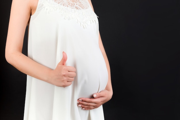 Ritagliata colpo di donna incinta positiva in abito bianco che mostra pollice in su gesto fresco sopra il suo pancione a sfondo nero. Buona maternità. Copia spazio.