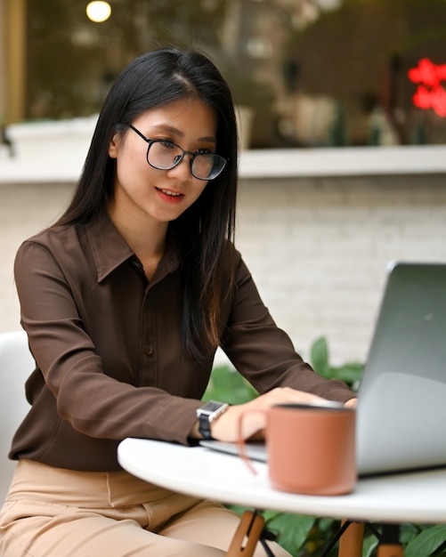 Ritagliata colpo di impiegato femminile con gli occhiali che lavora con il computer portatile sulla tavola rotonda nella caffetteria