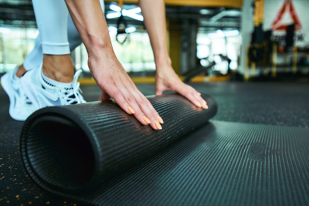 Ritagliata colpo di una donna fitness in abbigliamento sportivo che rotola tappetino yoga mentre si lavora in palestra. Sport, benessere e stile di vita sano