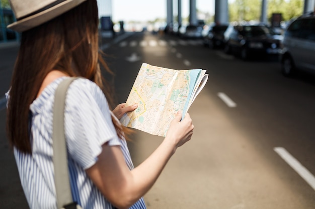 Ritagliata giovane turista turista con cappello con zaino alla ricerca del percorso nella mappa cartacea all'aeroporto internazionale