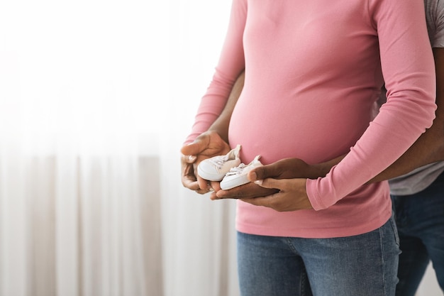 Ritagliato di irriconoscibile famiglia nera in attesa in casual tenendo insieme piccole scarpe per bambini. Uomo afroamericano e donna incinta con stivaletti per bambini in piedi vicino alla finestra a casa, primo piano, spazio per la copia