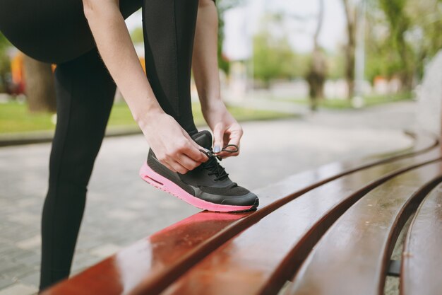 Ritagliato primo piano delle mani della donna che legano i lacci delle scarpe su scarpe da ginnastica femminili nere e rosa in allenamento su panchina all'aperto