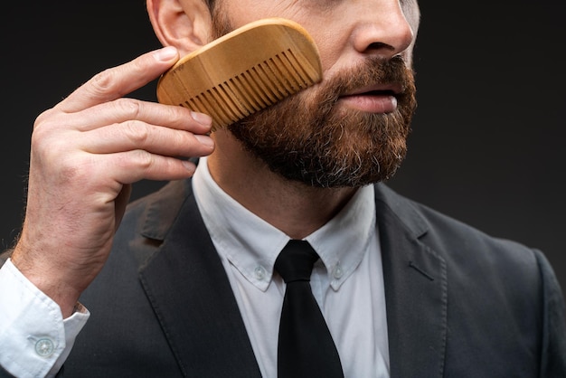 Ritagliato ritratto di uomo serio elegante con bella barba. Uomo che indossa una tuta che tiene il pettine mentre si trova da solo in uno studio su uno sfondo nero