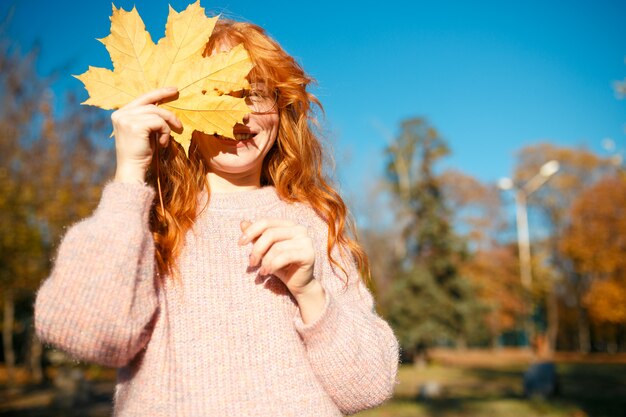 Ritratti di un'affascinante ragazza dai capelli rossi con un viso carino. Ragazza che posa nel parco di autunno in un maglione e una gonna color corallo. Nelle mani di una ragazza una foglia gialla